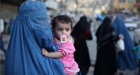 زن با چادر آبی و طفل در آغوش در جاده شلوغ